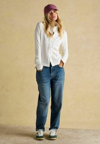 Weißes Hemd mit Spitzenakzenten, kombiniert mit lockeren Denim-Jeans und sneakers mit grünen Akzenten. Getoppt mit einer lila Mütze.