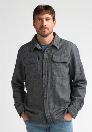 Mann mit leichtem Bart, trägt ein graues gemustertes Hemd mit Brusttaschen und blaue Jeans, steht mit den Händen in den Taschen.