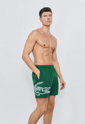 Grønne badeshorts med elastisk talje, sidelommer og en stor hvid krokodillegrafik på venstre ben. Glat tekstur.
