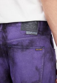 Paarse spijkerbroek met een zwart gestructureerd label en oranje "VOLCOM" borduring. De stof heeft een tie-dye patroon en een relaxte pasvorm.