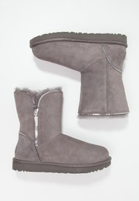 Šedé semišové kotníkové boty se zipem, měkkou kožešinovou podšívkou a texturovanou gumovou podrážkou. Obsahují lesklé detaily podél zipu.