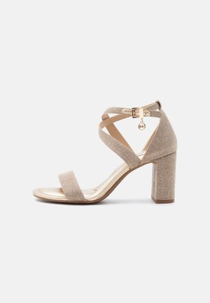 Μπεζ πέδιλο με ανοιχτά δάχτυλα και ψηλό τακούνι τύπου block, με λουράκια που σταυρώνονται στον αστράγαλο, χρυσή αγκράφα και ένα μικρό στρογγυλό γούρι κρεμασμένο από την αγκράφα.