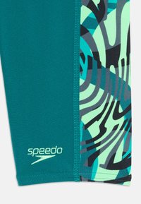 Turkosa badbyxor med ett kontrasterande grönt och svart abstrakt mönster på vänster sida. "speedo" logotyp tryckt längst ner.