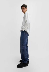 Felpa bianca, jeans blu scuro, scarpe nere. La felpa ha un collo tondo e maniche lunghe; i jeans hanno una gamba dritta e cuciture visibili.