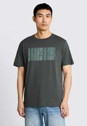 Mann trägt ein dunkelgraues T-Shirt mit vertikal gestreiftem Textdesign und hellblauen Jeans, steht vor einem schlichten hellen Hintergrund.