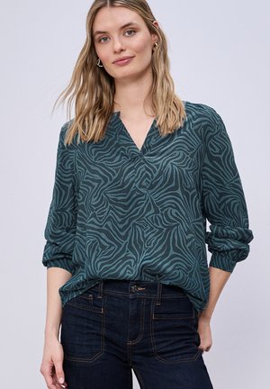 Jonge vrouw die een lange mouw blouse met een teal en zwarte zebra print draagt, gecombineerd met donkerblauwe jeans, staand met één hand in de zak tegen een grijze achtergrond.