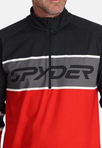 Pullover a mezza zip in nero, grigio e rosso. Presenta un grande logo "SPYDER" sul petto e strisce a contrasto. Realizzato in materiale morbido ed elastico.