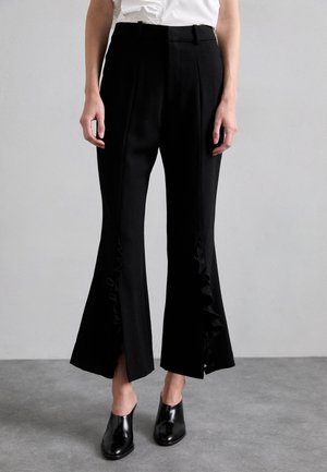 Black flared trousers with ruffled side openings, featuring a smooth fabric, high waist, and side pockets. Paired with black mules.