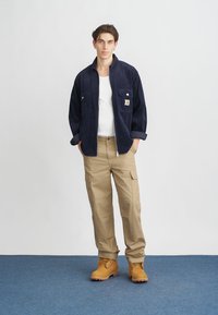 Marine corduroy jack met twee voorzakken, een wit t-shirt eronder, tan cargobroek en bruine veterschoenen. Blauwe tapijten achtergrond.