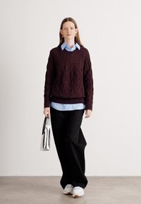 Raudonos vyno spalvos megztinis su tekstūrine struktūra, dėvimas ant šviesiai mėlynos spalvos marškinių su apykakle. Juodos plačios kelnės ir sidabrinė rankinė papildo įvaizdį.