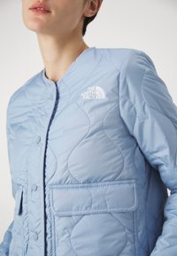 Jasnoniebieska pikowana kurtka o zaokrąglonych kształtach, z zapięciem na zatrzaski z przodu i dwoma naszywanymi kieszeniami. Logo The North Face znajduje się na lewej piersi.