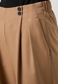 Béžové plisované kalhoty s hladkou texturou, dvě tmavé knoflíky v pase, elastický pás a detail boční kapsy.