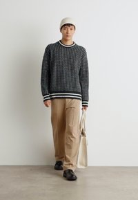 Suéter de patrón en blanco y negro con puños acanalados, pantalones beige, zapatos negros y un gorro crema, sosteniendo una bolsa de mano de color claro.