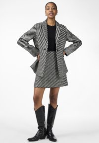 Rutet blazer- og skjørtsett i svart og hvitt med teksturert stoff. Fullført med svarte knelangstøvler. Skreddersydd passform med markante revers.