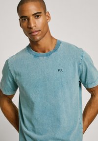 Světle modré denimové tričko s krátkými rukávy, kulatým výstřihem a malým námořnickým logem na hrudi. Měkká textura a volný střih.