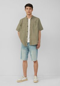 Korte mouw, olijfgroene overhemd met knopen en twee borstzakken, gecombineerd met lichtblauwe denim shorts en witte sneakers met beige accenten.