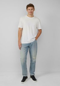 Biała koszulka z okrągłym dekoltem, jasnoniebieskie dżinsy typu straight-leg z przetarciem na kolanie, czarne sneakersy z białą podeszwą, stojąca na neutralnym tle.