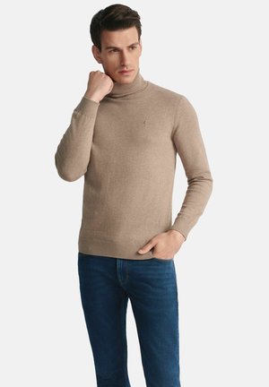 Beżowy sweter typu golf z prążkowanymi mankietami i dołem, z małym logo na piersi. W zestawie z ciemnoniebieskimi dżinsami.