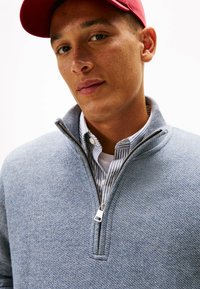 Maglione azzurro chiaro con cerniera a tutta lunghezza e lavorazione a maglia testurizzata, con collo e camicia bianca a strisce blu sotto. Cappellino rosso indossato sopra.