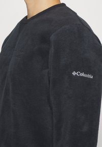 Mustavillainen fleece-huppari, jossa on V-kaula-aukko, teksturoitu kangas ja brodeerattu Columbia-logo vasemmassa hihassa.