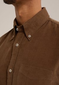 Camisa de pana marrón con cuello abotonado, bolsillo delantero y una sutil textura de canalé; abotonada con botones de color claro.