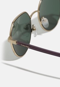 Ochelari de soare Calvin Klein cu lentile verzi unghiulare, cadru din metal auriu și brate din plastic violet cu detalii gravate ale logo-ului.