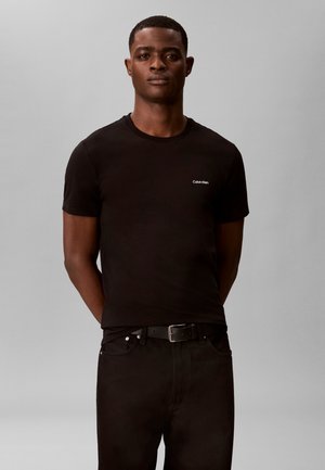 Homme portant un t-shirt noir Calvin Klein et un pantalon noir avec une ceinture, debout devant un fond uni gris clair.