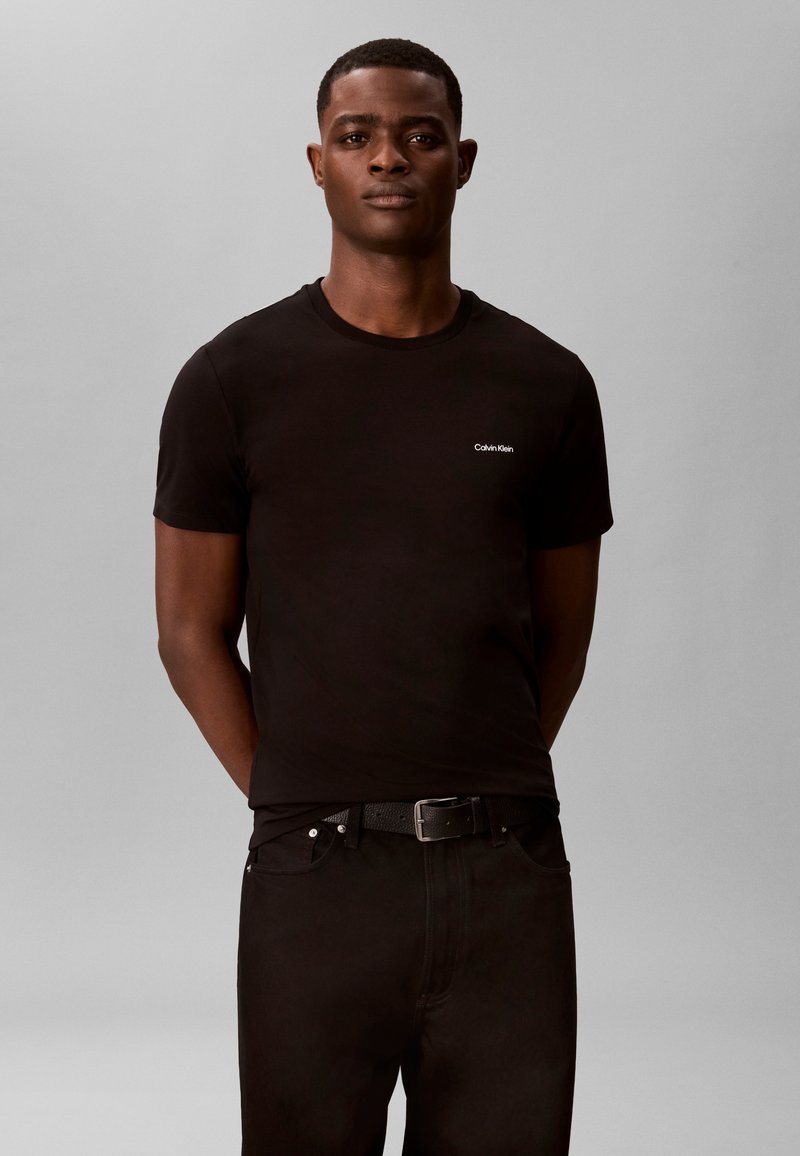 Άνδρας που φοράει μαύρη μπλούζα Calvin Klein και μαύρο παντελόνι με ζώνη, στέκεται μπροστά σε απλό ανοιχτό γκρι φόντο.