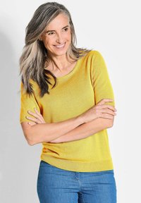 Gelber Strickpullover mit kurzen Ärmeln und rundem Ausschnitt, kombiniert mit blauen Jeans. Der Stoff wirkt weich mit einer glatten Textur und einem subtilen Glanz.