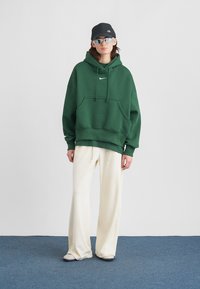 Grønn hettegenser med lomme foran, hvit Nike-logo. Kombinert med kremfargede vide bukser og en svart caps, iført reflekterende solbriller.