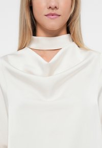 Hvid satin top med høj hals og en udskæringsdetalje. Har en glat tekstur og et draperet halsdesign.