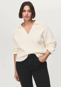 White, ribbed knit sweater with a V-neck and fold-over collar; worn over black jeans. The sleeves are rolled up for a relaxed fit.