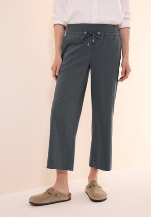 Pantaloni grigi alla caviglia con cintura elastica e cordoncino regolabile; dotati di tasche laterali e una vestibilità rilassata. Abbinati a zoccoli color nocciola.