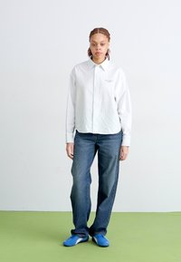 Camisa blanca de mangas largas, con cuello y rayas, sobre jeans azul oscuro, acompañada de zapatos azules. Diseño simple sin logotipos ni patrones visibles.
