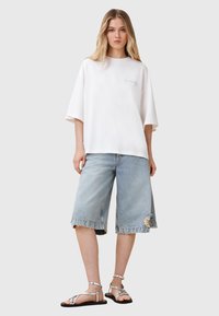 Biały oversize'owy t-shirt z logo, w zestawieniu z podartymi jasnoniebieskimi szortami dżinsowymi oraz srebrnymi sandałami na paski. Prosty i casualowy design.