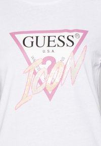 Hvid T-shirt med et centralt pink trekantet logo, som har teksten "GUESS U.S.A." og en glitrende "ICON" i kursiv skrifttype.
