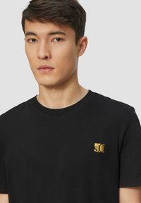 Schwarzes Baumwoll-T-Shirt mit rundem Halsausschnitt, ausgestattet mit einem kleinen Gold-Logo-Patch auf der linken Seite, glatte Textur und kurzen Ärmeln.