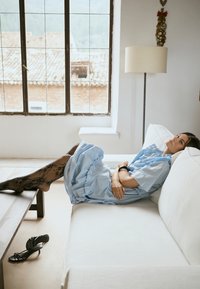 Ženska v modri obleki in črnih vzorčanih najlonkah se zleknjena na belem kavču z nogami na lesenem mizi, črni čevelji z visoko peto so na tleh v bližini.