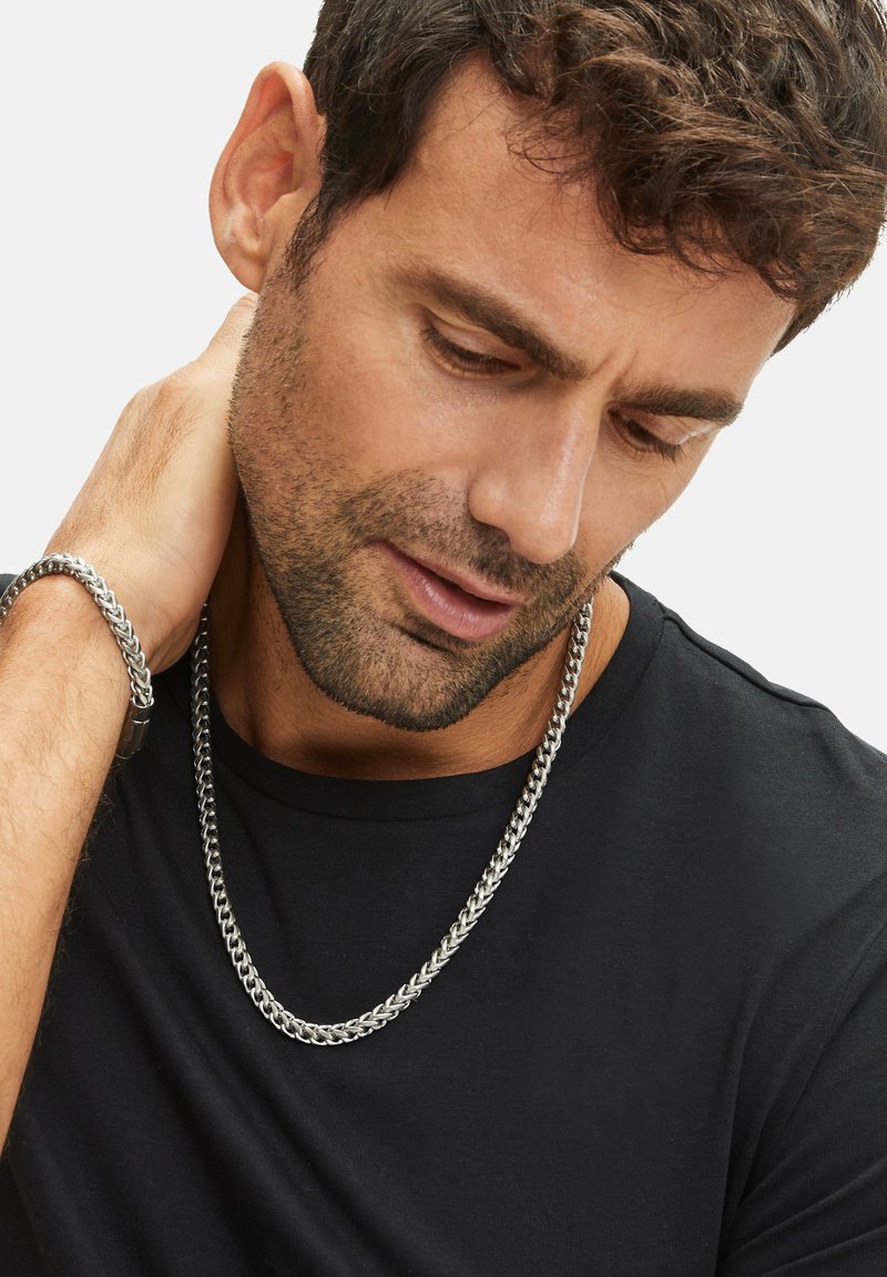 Zilveren ketting, dikke schakels, gedragen over een zwart t-shirt. Armband past bij de ketting, gladde textuur, lus ontwerp, reflecterende afwerking.