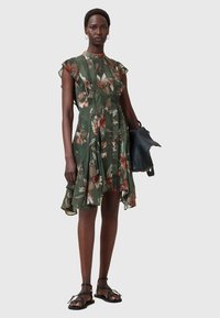 Floral green dress with short ruffled sleeves, pleated design, asymmetrical hem, worn with black sandals and a black handbag.