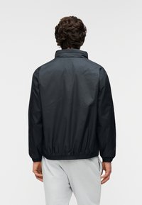 Ελαφρύ μαύρο μπουφάν με ψηλό γιακά, ελαστικές μανσέτες και μέση, με λεία υφή και χωρίς ορατά λογότυπα ή σχέδια.