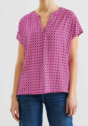 Kvinde iført en lyserød bluse med korte ærmer og et geometrisk cirkelmønster samt mørkeblå jeans, stående foran en ensfarvet baggrund.