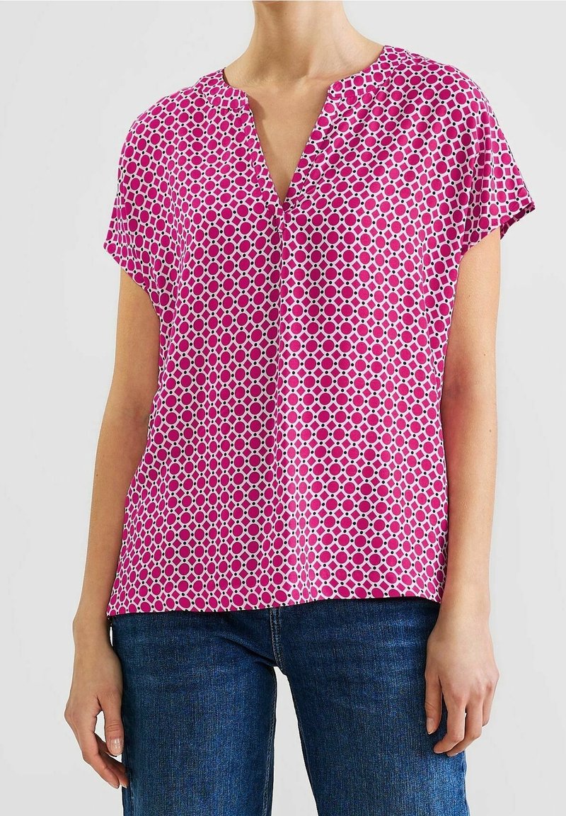 Femme portant un chemisier rose à manches courtes avec un motif géométrique de cercles et un jean bleu foncé, debout devant un fond uni.