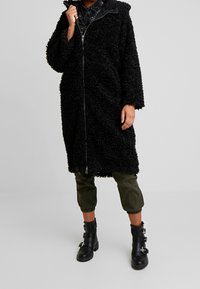 Abrigo negro de peluche con cierre frontal, grandes bolsillos y capucha. Lleva puesto encima de pantalones cortos de color verde oliva y botas de combate negras con correas de hebilla.