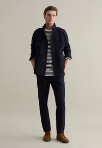 Marineblaues Sakko mit zwei Brusttaschen über einem weiß-blau gestreiften Hemd, kombiniert mit marineblauen Hosen und braunen Wildlederstiefeln. Schlichte, legere Kleidung.