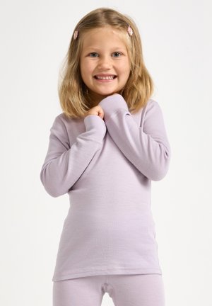 Jong meisje met schouderlang blond haar en roze haarspeldjes, glimlachend, gekleed in een lichtpaarse top met lange mouwen en bijpassende legging, handen dichtbij de borst.