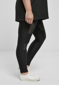 Czarne, prążkowane legginsy z przezroczystymi panelami z siateczki, w połączeniu z luźną czarną bluzą i białymi trampkami, podkreślające dopasowany krój i teksturę.