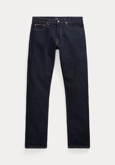 Στενά τζιν σκούρου μπλε χρώματος με πέντε τσέπες, χάλκινες τάπες και ορατές πορτοκαλί ραφές, τοποθετημένα επίπεδα σε φωτεινό φόντο.