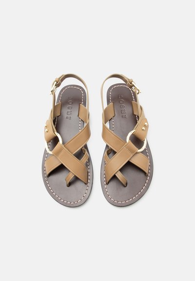 Καφέ δερμάτινα σανδάλια με επίπεδη σόλα και σταυρωτούς ιμάντες, με χρυσό δαχτυλίδι και αγκράφες στον αστράγαλο πάνω σε σκούρες σόλες με λευκή ραφή.