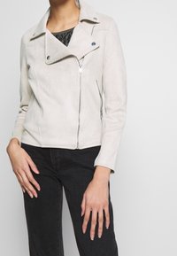 Motocyklowa kurtka z jasnoszarego sztucznego zamszu z zamkiem błyskawicznym, kołnierzem na zatrzask i skośnymi kieszeniami. Charakteryzuje się dopasowanym krojem i gładką fakturą.